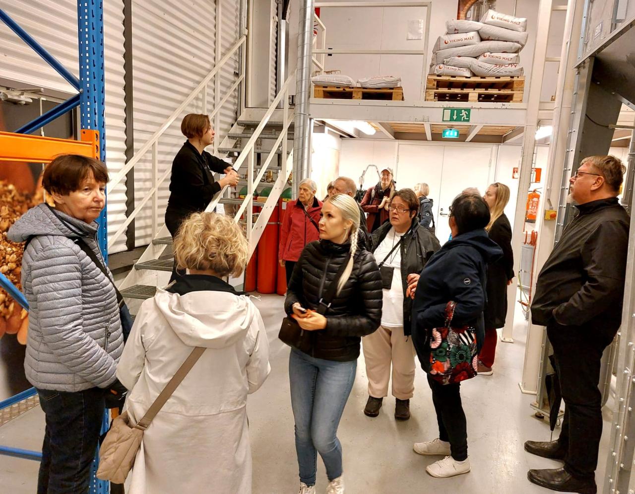 Tuotantotilojen aulassa, jossa vasemmalla esittelijä seisoo metallikaiteisilla portailla. Osa ihmistä katsoo häntä ja osa tutkailee ympärilleen.